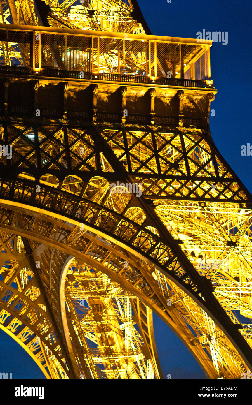 Eiffelturm beleuchtete Eisenstruktur Detail Goldene Lichter bei Nacht Paris Frankreich // PARIS, Frankreich - Detail der ikonischen schmiedeeisernen Struktur des Eiffelturms wird mit goldenen Lichtern vor dem Nachthimmel beleuchtet. Die Nahaufnahme zeigt das komplizierte Gitterwerk und die Namen von 72 französischen Wissenschaftlern, Ingenieuren und Mathematikern, die um die erste Ebene graviert sind. Dieses Wahrzeichen in Paris, das aus Pfütscheisen gebaut wurde, wurde von Gustave Eiffel für die Ausstellung Universelle 1889 gebaut. Die goldene Beleuchtung des Turms, die 1985 eingeführt wurde, wird durch eine funkelnde Lichtshow t ergänzt Stockfoto