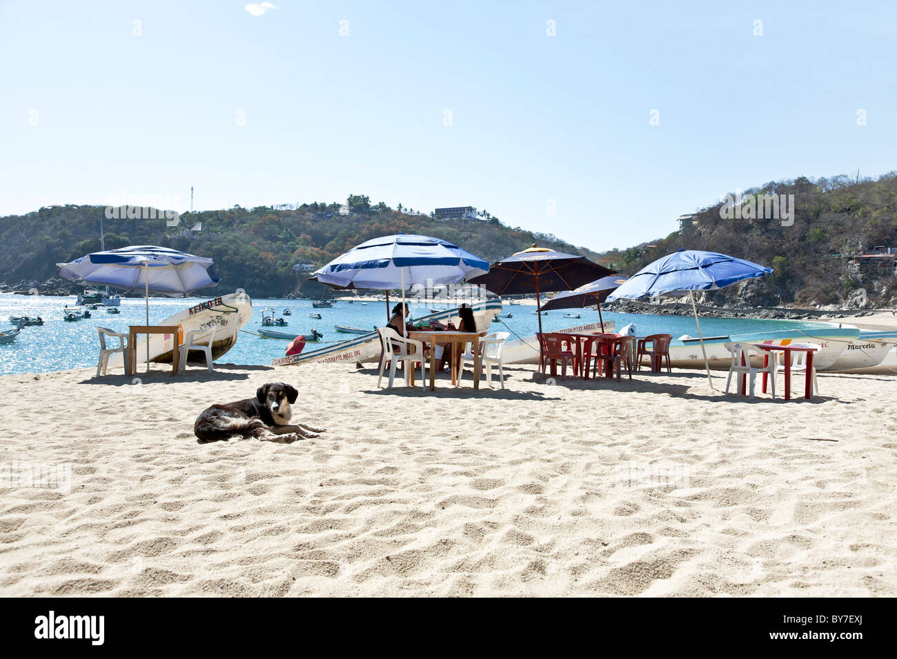 Clustered Sonnenschirme Schützen der Erfrischung Tische mit Blick auf kleine Boote im Wasser azurblauen Meer und den Hügeln, die das Dorf Mexiko Puerto Angel Stockfoto