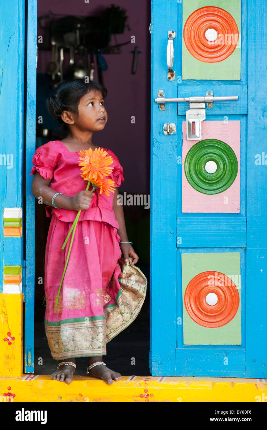 Jungen Säugling Inderin orange Gerbera Blume Stand in der Tür ihres bunten Hauses halten. Andhra Pradesh, Indien Stockfoto