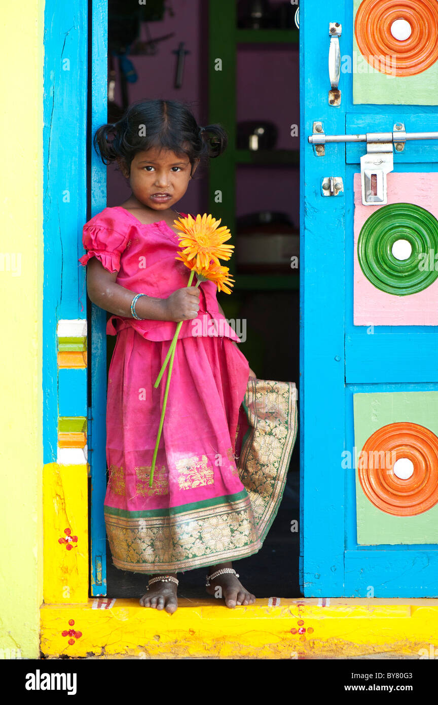 Jungen Säugling Inderin orange Gerbera Blume Stand in der Tür ihres bunten Hauses halten. Andhra Pradesh, Indien Stockfoto