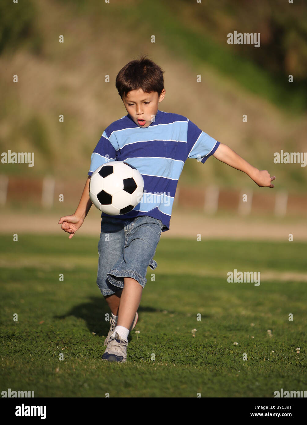 Authentische glücklich Latino Boy spielt mit Fussball Ball im Feld tragen blau gestreiften t-Shirt. Stockfoto
