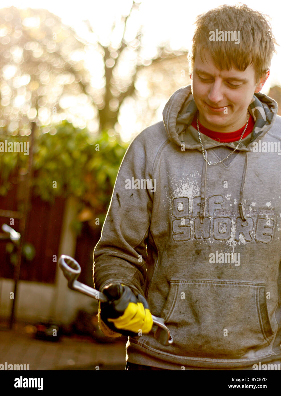 Einen jungen kaukasischen männlichen Mechaniker greifen einen Schraubenschlüssel, trägt ein paar schützende Rigger Handschuhe und ein Hoodie. Stockfoto