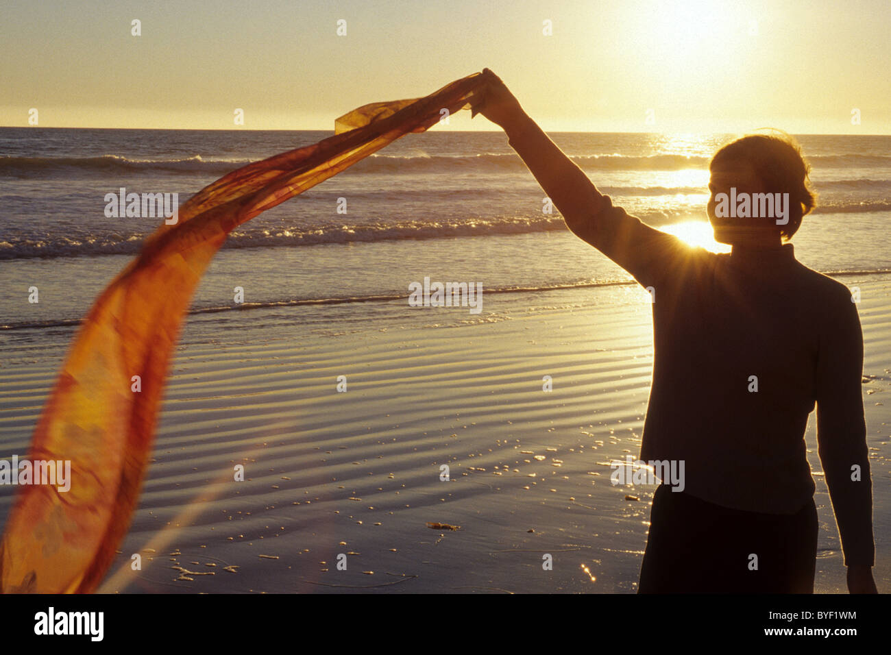 Frau in der Silhouette hält einen Schal am Strand bei Sonnenuntergang. Stockfoto