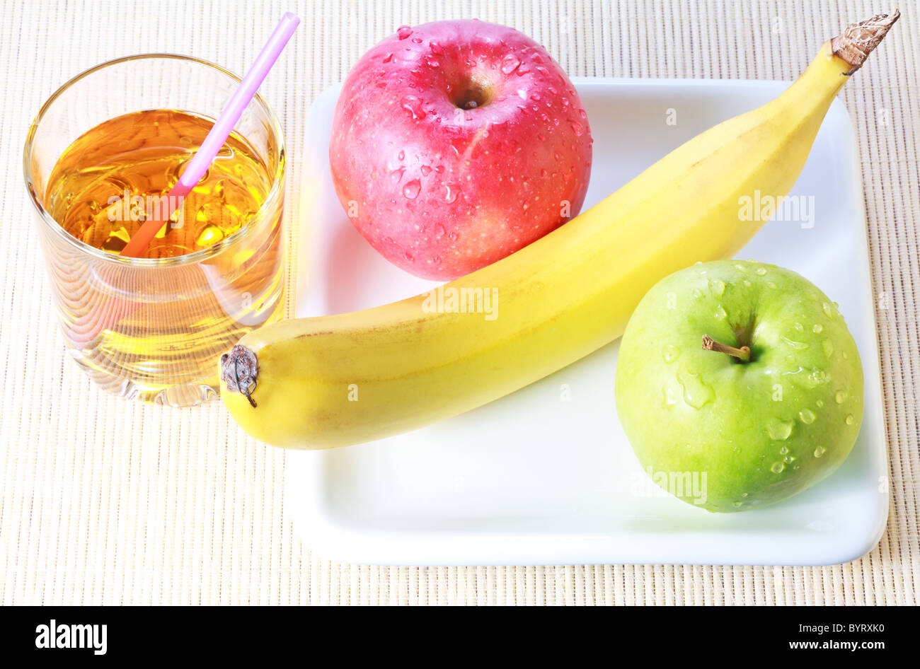 Banane, grüne und rote Äpfel auf weißen Teller und Glas Apfelsaft über Bambus Matte Hintergrund Stockfoto