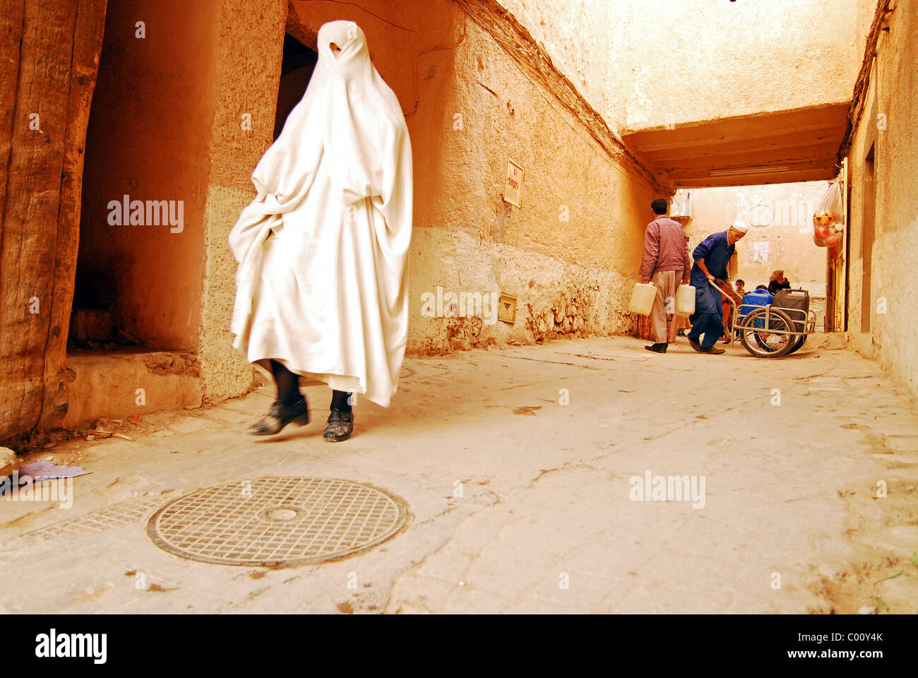 Algerien, Ghardaia, traditionell gekleidete Frau im Schleier zu Fuß auf schmalen Durchgang umgeben von Mauern, Männer halten Behälter Stockfoto