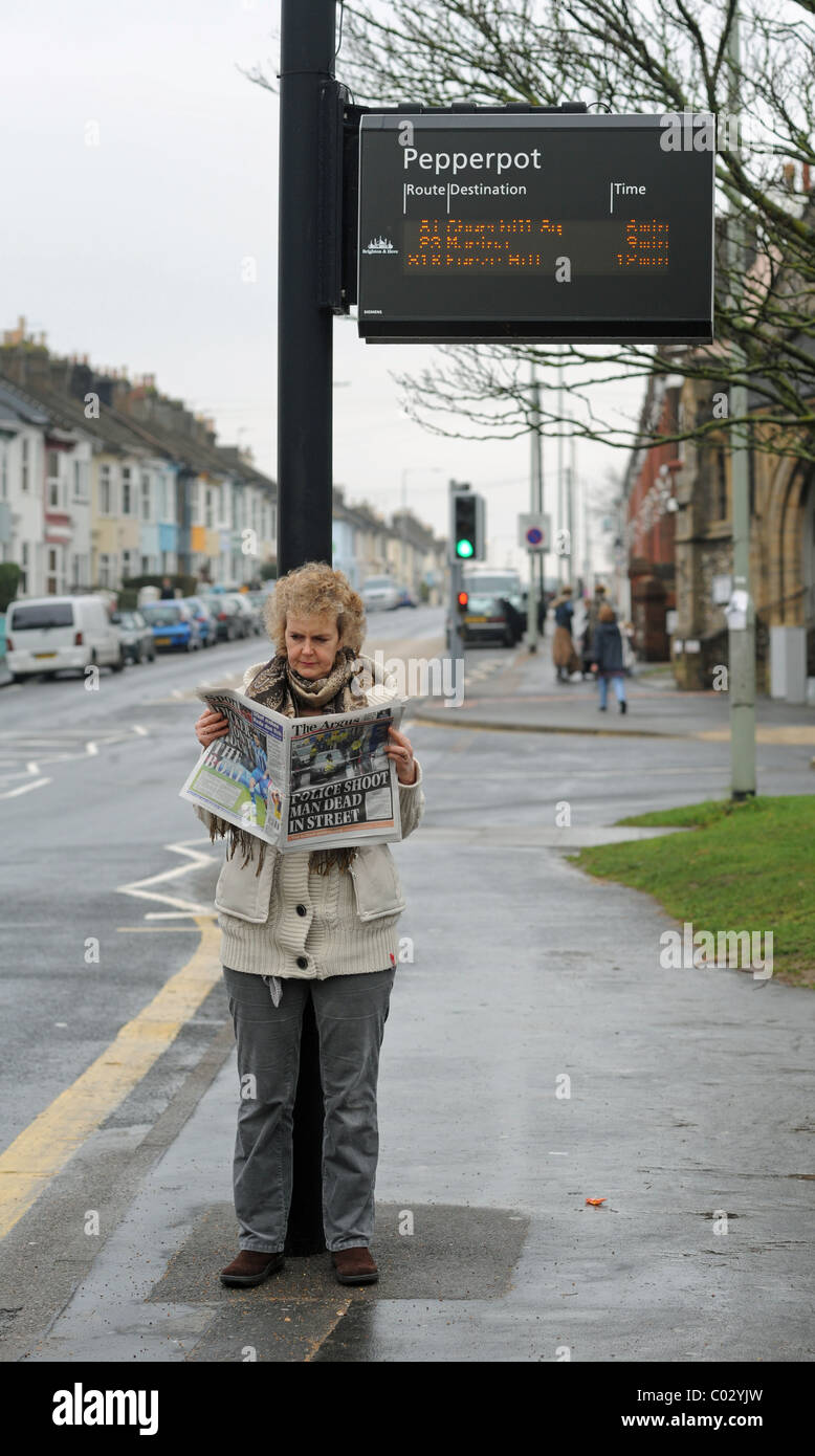 Frau liest Lokalzeitung unter einem Bus Fahrplan Hinweisschild Brighton UK Stockfoto