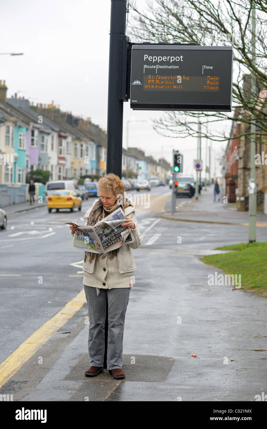 Frau liest Lokalzeitung unter einem Bus Fahrplan Hinweisschild Brighton UK Stockfoto