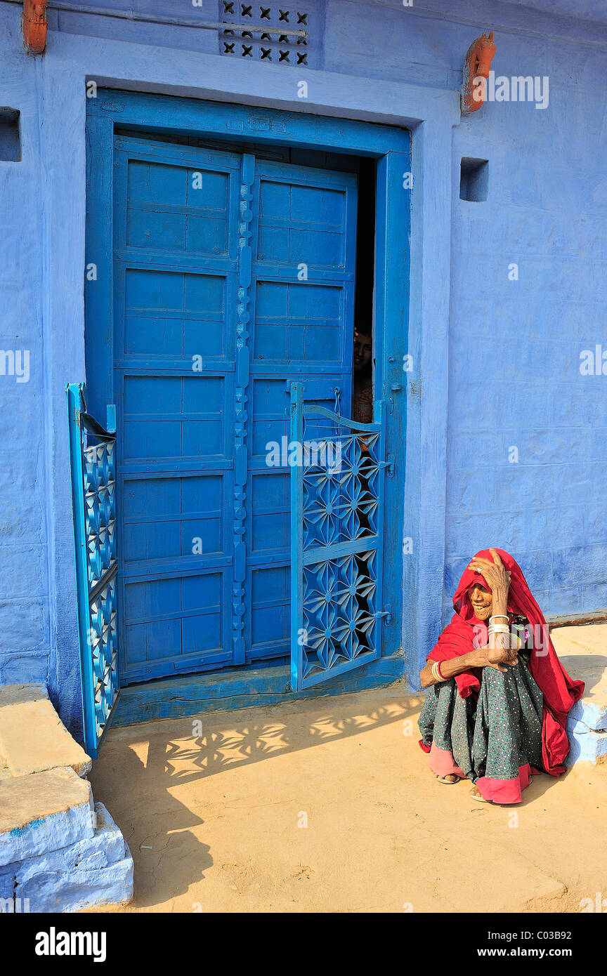 Ältere indische Frau trägt einen roten Sari sitzen auf dem Boden vor einer blau lackierten Tür, Thar-Wüste, Rajasthan, Indien Stockfoto