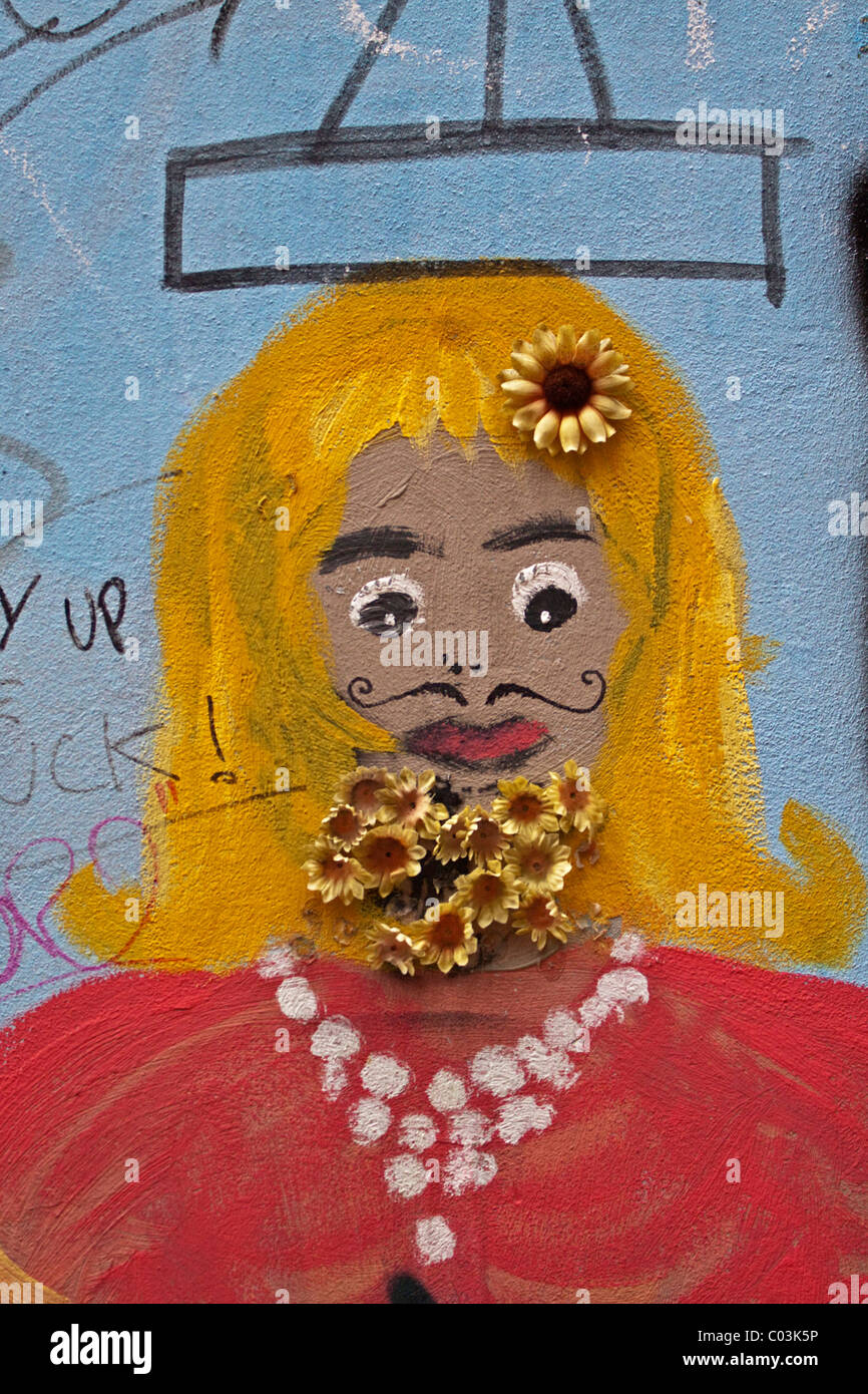 Bemaltem Gesicht mit blonden Haaren an einer Wand mit künstlichen Blumen Stockfoto