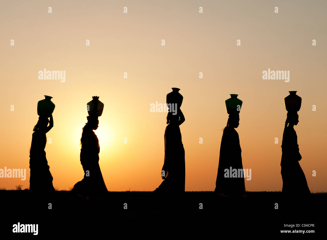 Gruppe von indischen Frauen auf ihren Köpfen in der indischen Landschaft bei Sonnenuntergang mit Wasser Töpfe wandern. Silhouette Stockfoto