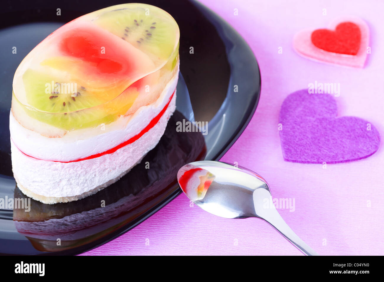 Nahaufnahme von Kuchen mit Gelee und Früchte auf einem schwarzen Teller mit Valentinstag Herz geformt Dekorationen auf lila Hintergrund Stockfoto