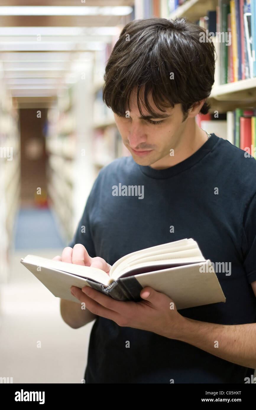 Ein junger Mann liest ein Buch in der Bibliothek stehen in den Gängen der Regale. Stockfoto