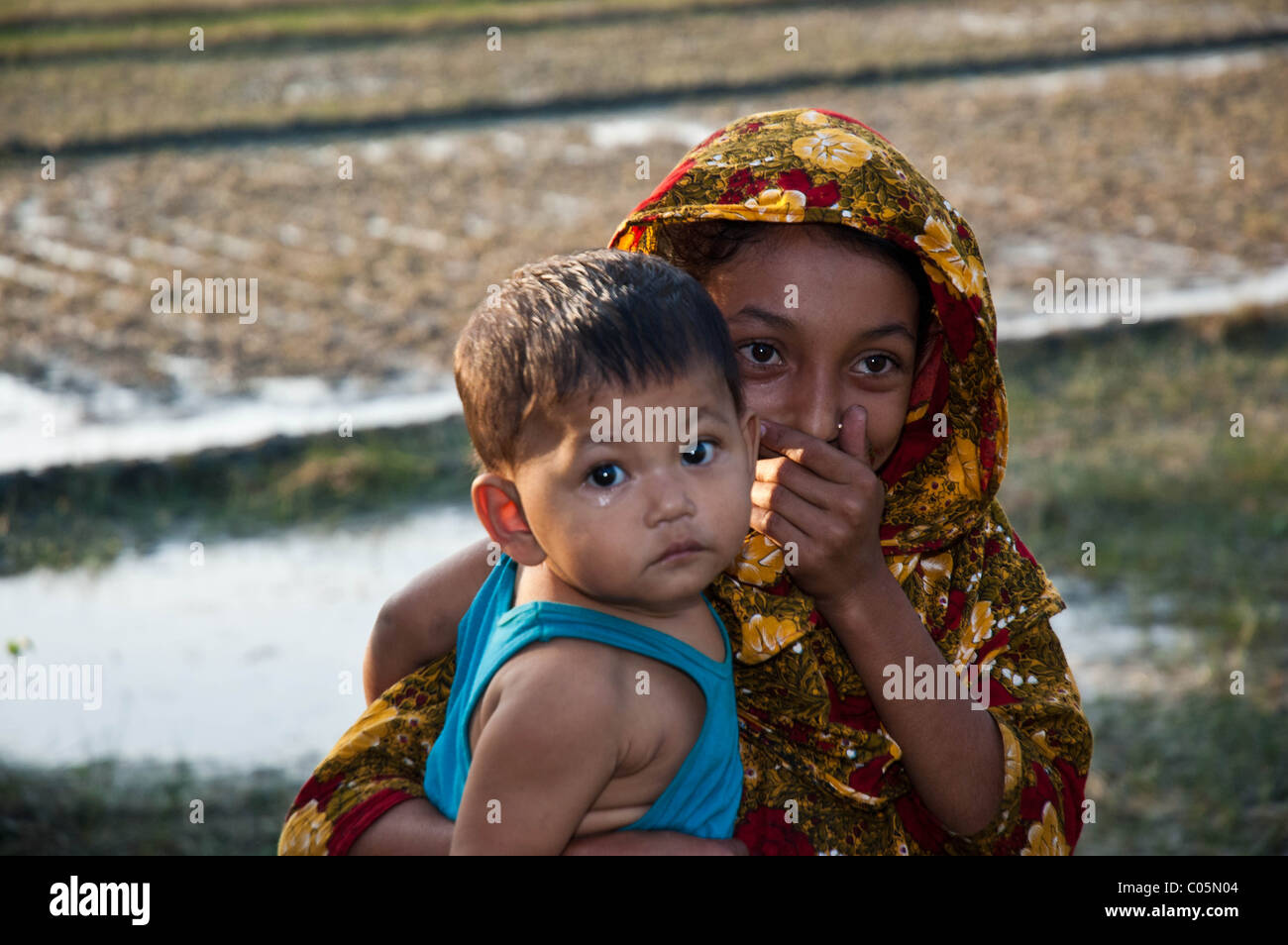 Eine junge Mädchen aus Bangladesch in einem Reisfeld, ihr jüngeren Bruder in einem Arm, während den Mund mit ihr auf andere Hand hält Stockfoto