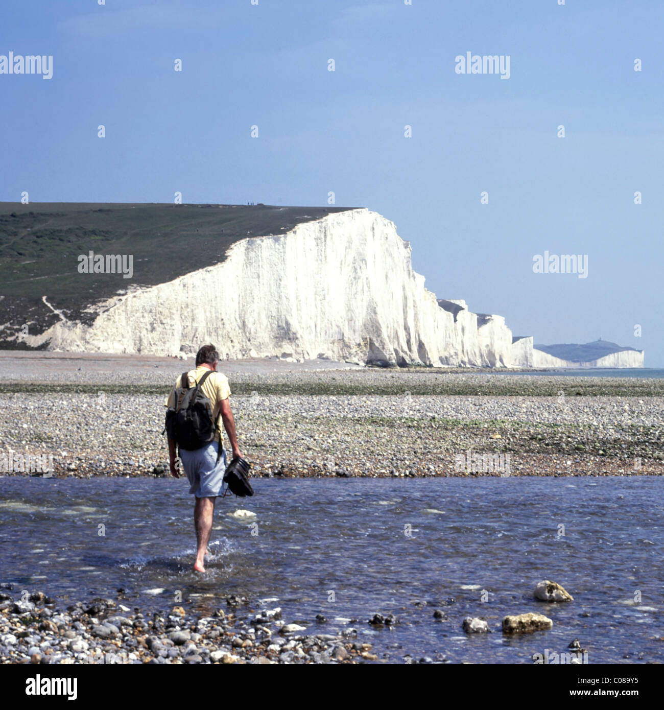River Cuckmere Haven Mündung Seven Sisters weiße Kreidefelsen mit einem Mann, der durch das Wasser geht und seine Stiefel hält, mit dem Rucksack East Sussex England UK Stockfoto
