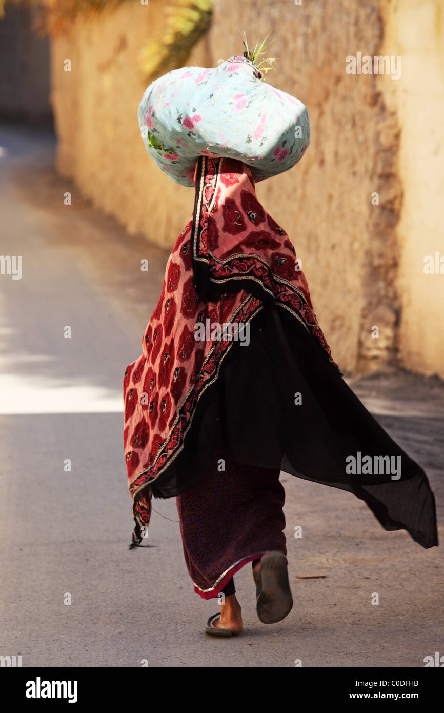 Eine omanische Frau trägt eine Tasche auf hören Kopf während des Gehens in einem Dorf. Stockfoto