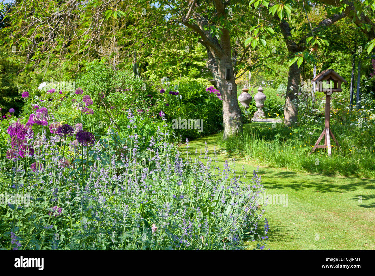 Eine Ecke eines englischen Gartens mit einem grasbewachsenen Weg zwischen Blumenrabatten und ein Büschel von schattigen Bäumen Stockfoto