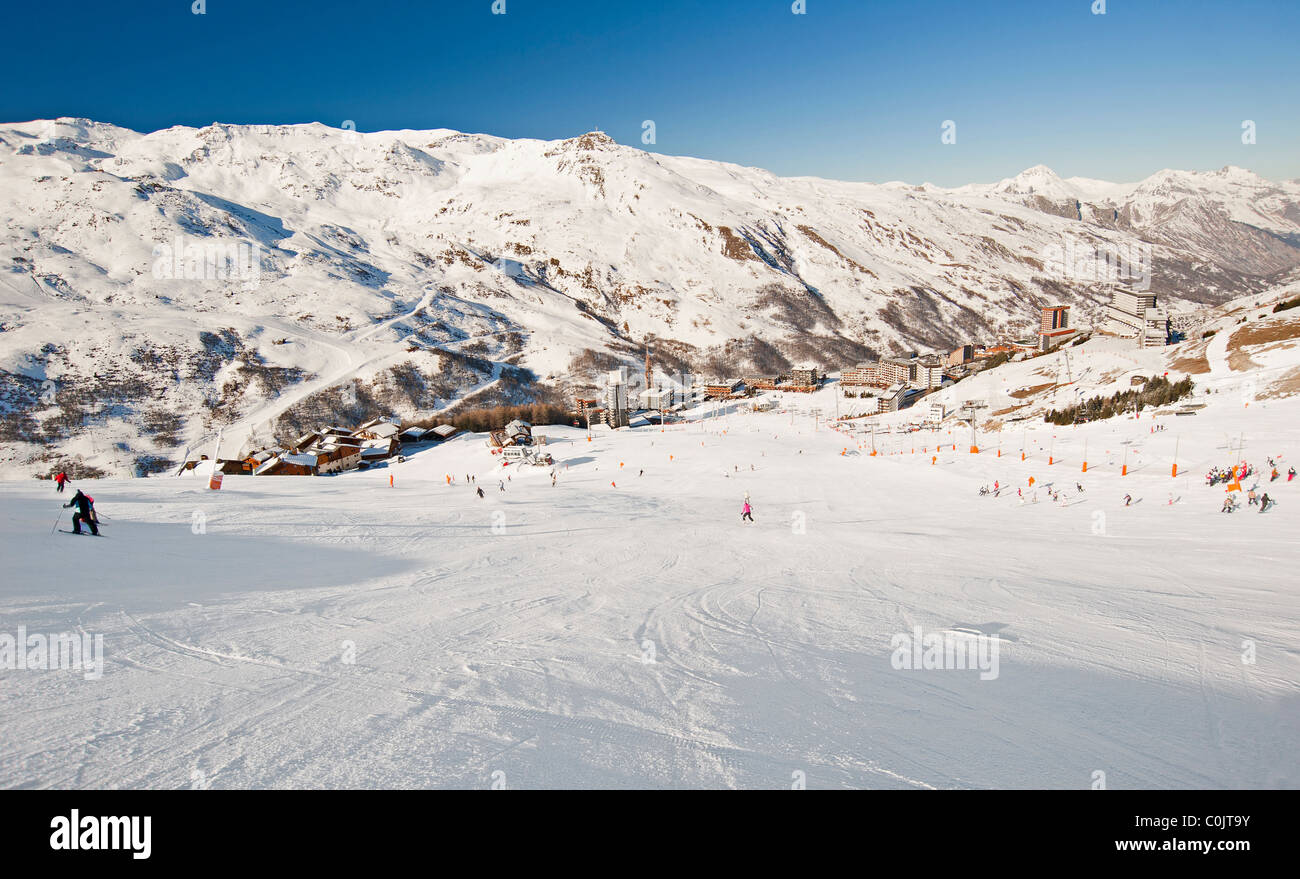 Zeigen Sie auf der Piste zu einem Dorf mit Skifahrer und Bergen im Hintergrund an Stockfoto