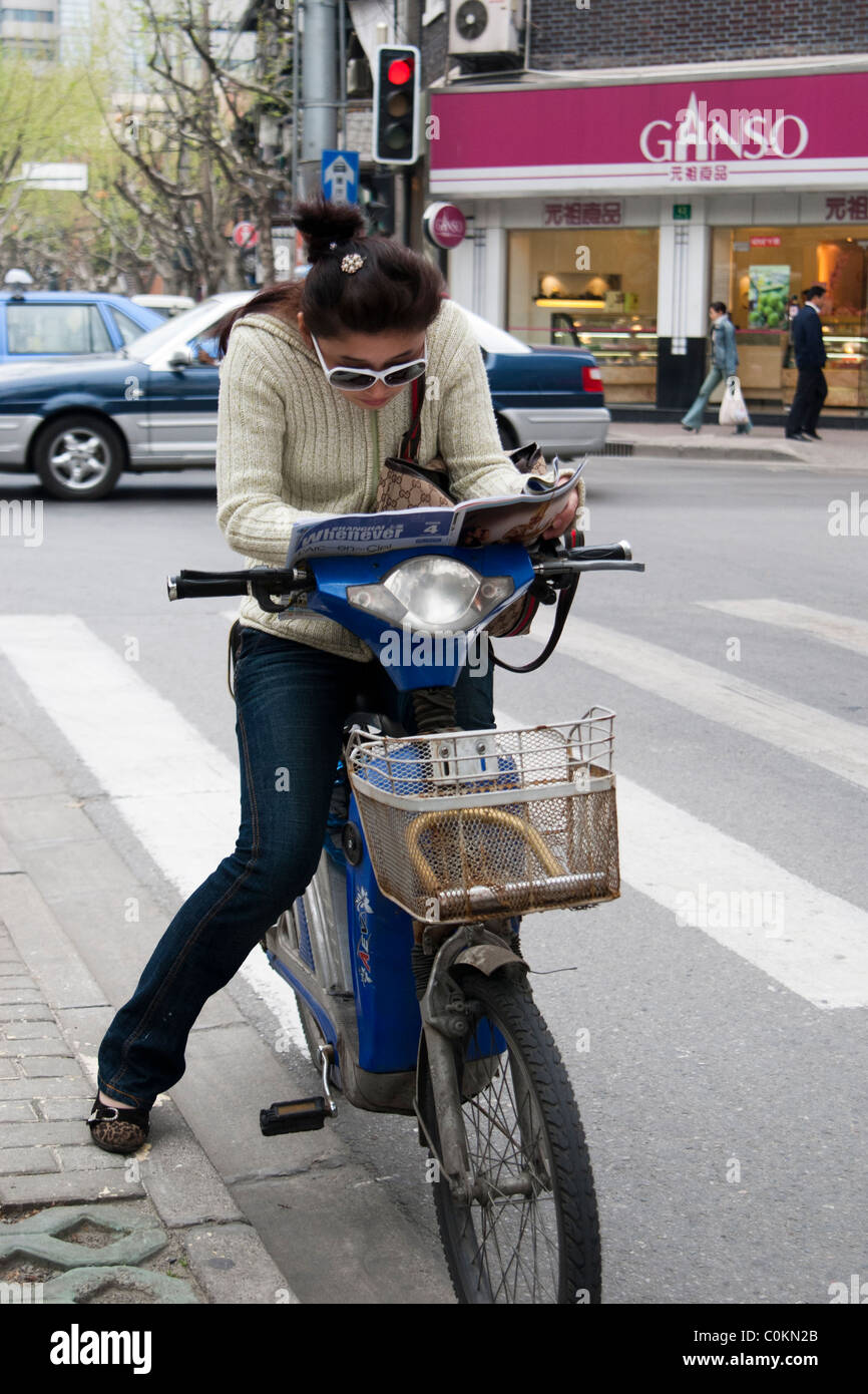 Chinesische Frau liest eine Zeitschrift in einer Straße beim Sitzen auf dem Fahrrad Stockfoto