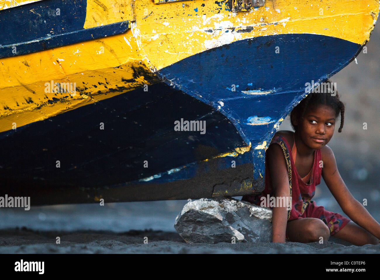 Ein junges Mädchen sitzt auf dem Strand neben einem bunten hölzernen Fischerboot in der Nähe von Sao Filipe, Fogo Island, Cape Verde Stockfoto