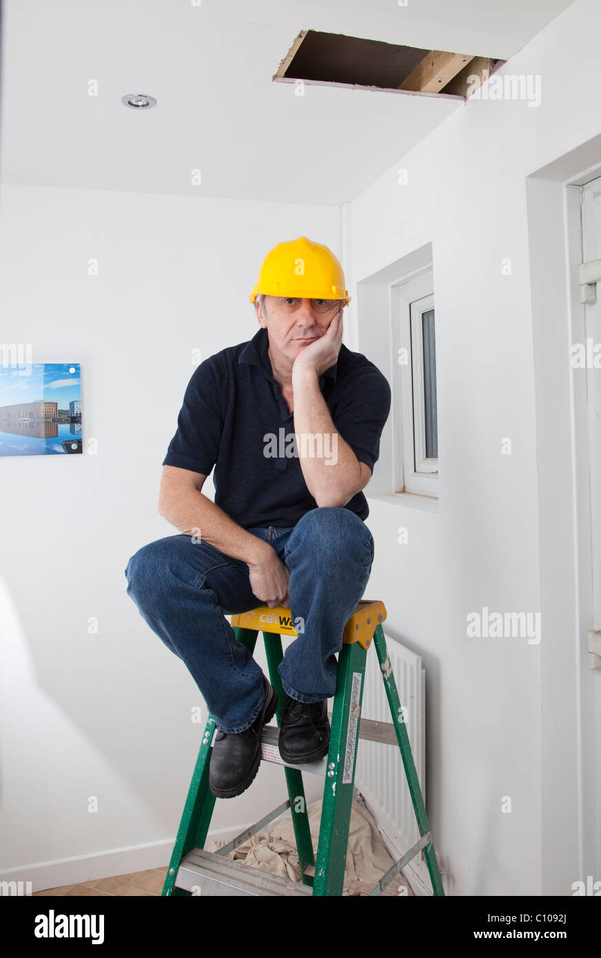 Mann mit gelben Sicherheit Hut sitzt auf Leiter unter einem Loch in der Decke Stockfoto