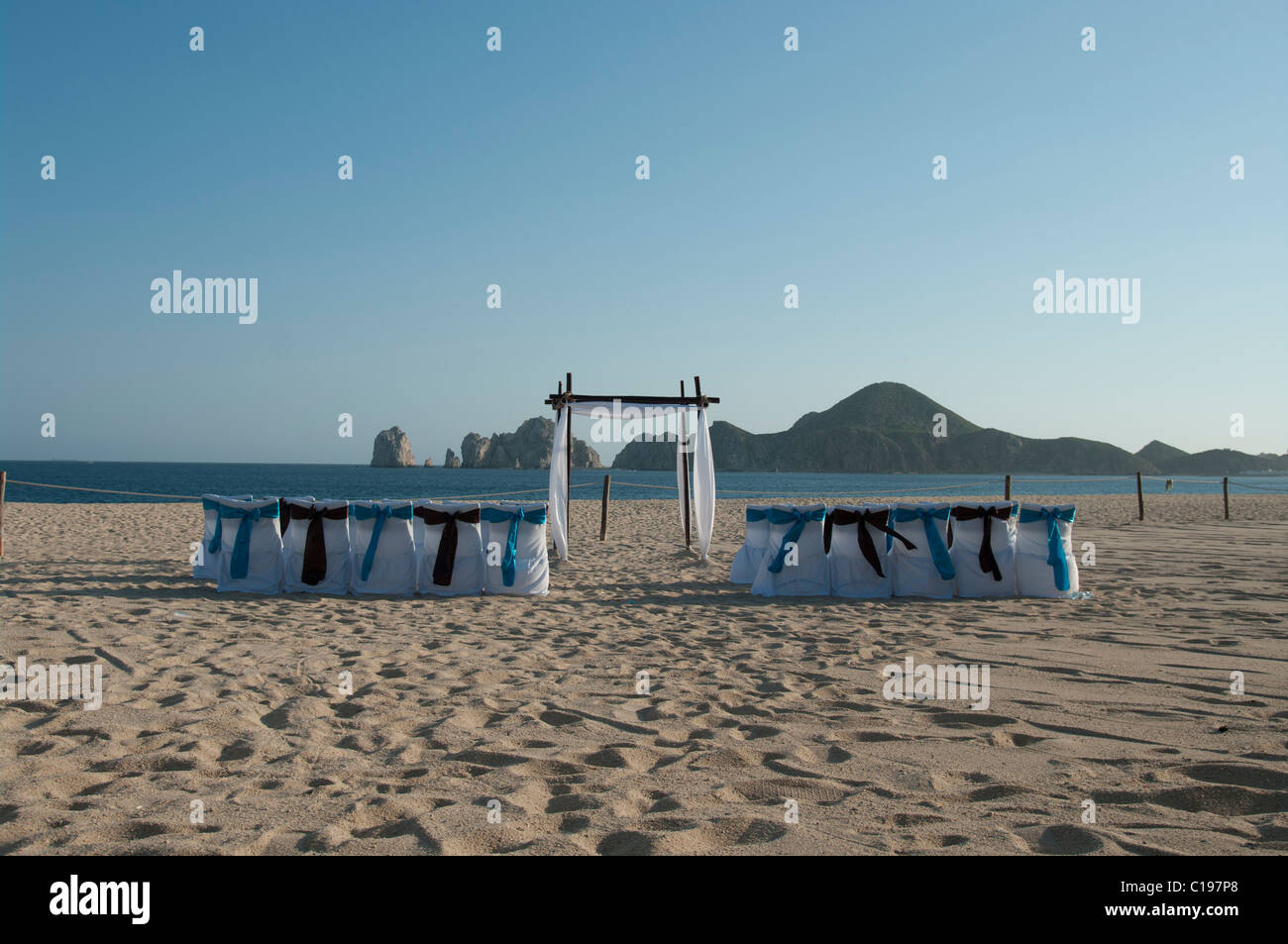 Ein verlassener Strand eingerichtet für eine Trauung.  Die Stuhlhussen sind weiß mit blauen und braunen Bögen. Ein Bogen erwartet das Paar. Stockfoto