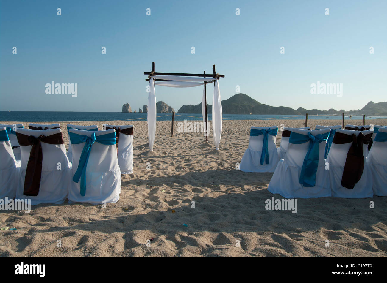 Ein verlassener Strand eingerichtet für eine Trauung.  Die Stuhlhussen sind weiß mit blauen und braunen Bögen. Ein Bogen erwartet das Paar. Stockfoto