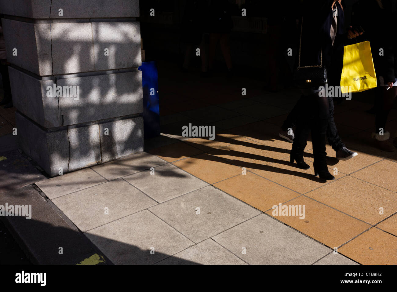 Eine Selfridges-Shopper mit dem eingebrannten gelben Beutel geht vorbei an die schattenhafte Ritz Worte auf einer Straße Säule. Stockfoto