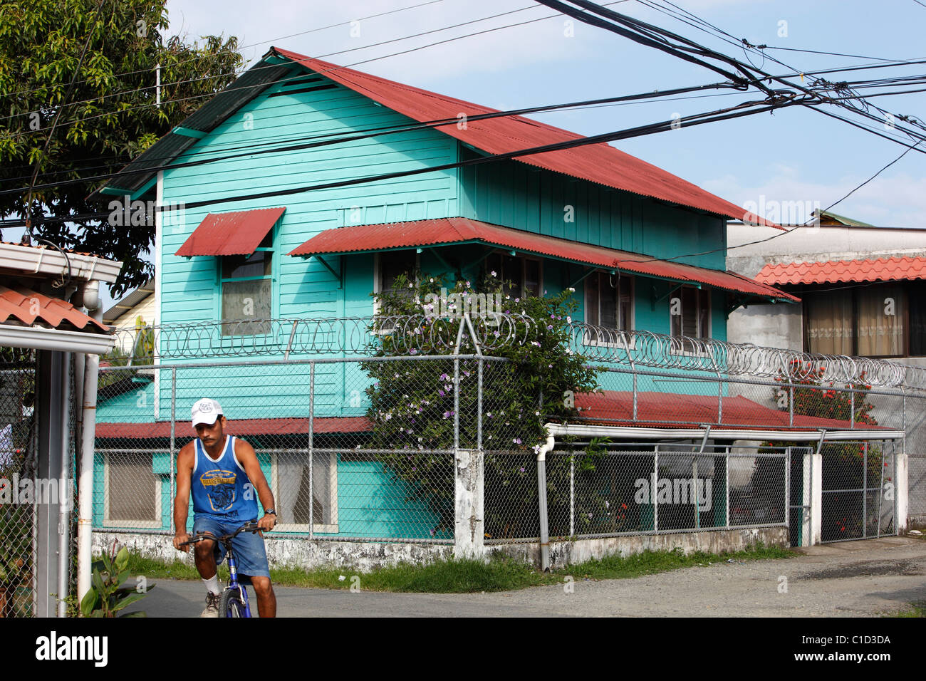 Ein Mann fährt Fahrrad vorbei an einem Haus umgeben von einem Zaun und Stacheldraht in einer Nachbarschaft von Quepos, Costa Rica Stockfoto
