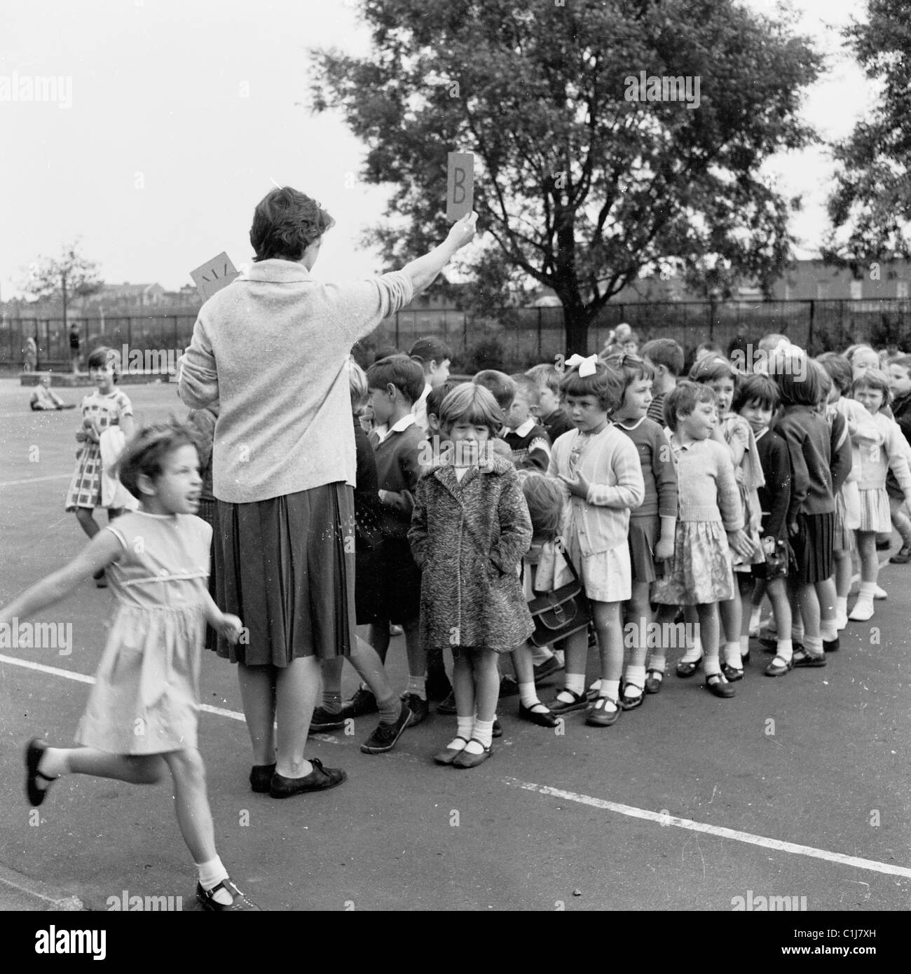 1960er Jahren Lehrerin hält ein Schild (Buchstabe B) in der rechten Hand, wie sie eine Gruppe von jungen Kindern eine Linie auf einem Schulhof organisiert. Stockfoto