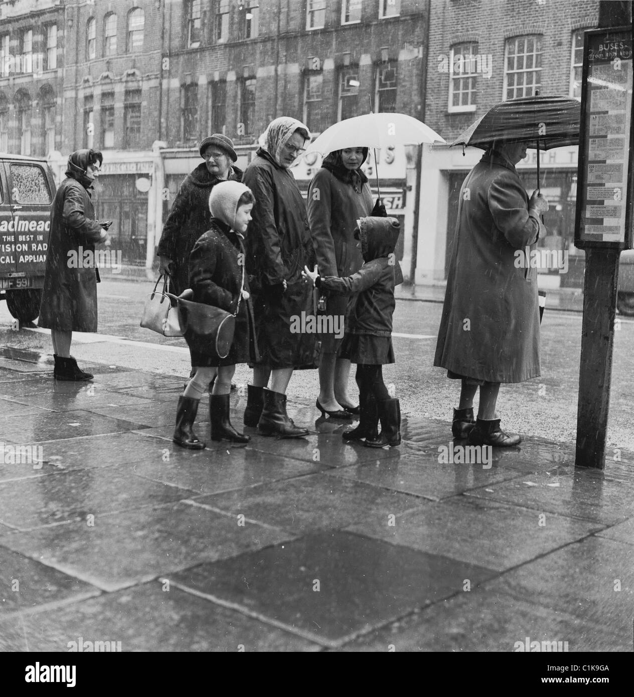 1960s, an einer Bushaltestelle, wartet eine Mutter mit ihren zwei kleinen Kindern mit anderen Damen auf einem Bürgersteig im Regen auf einen Bus, London, England, Großbritannien. Stockfoto