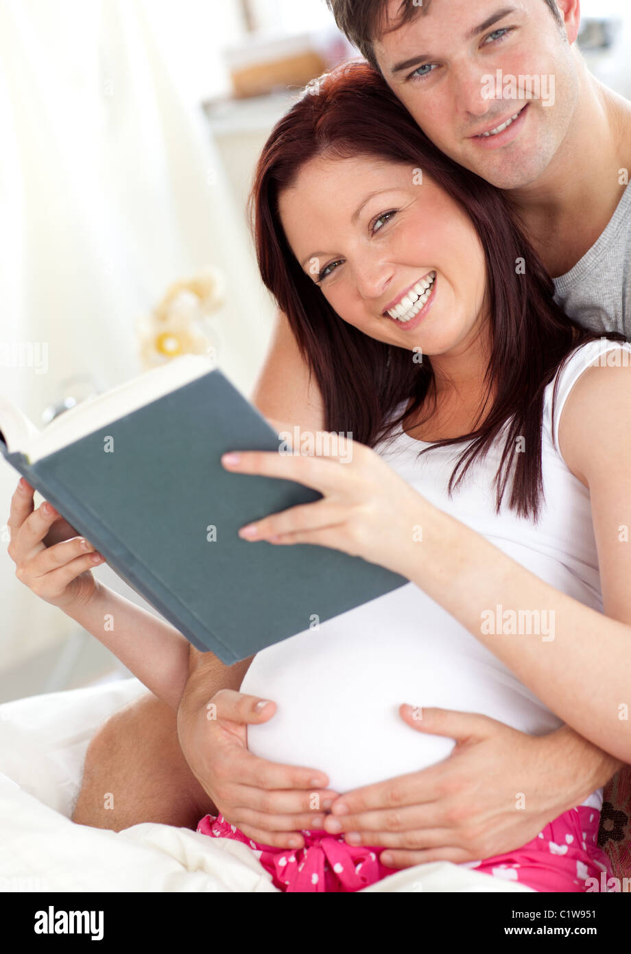 Fröhliche zukünftige Eltern Lesen eines Buches sitzt auf ihrem Bett im Laufe des Vormittags Stockfoto