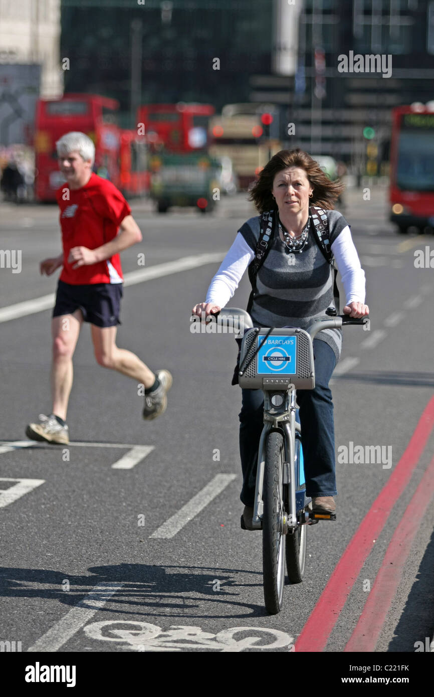 Eine weibliche Radfahrer mit dem Boris-Bike, in London, England, mit einem Mann auf der anderen Straßenseite im Hintergrund laufen Stockfoto