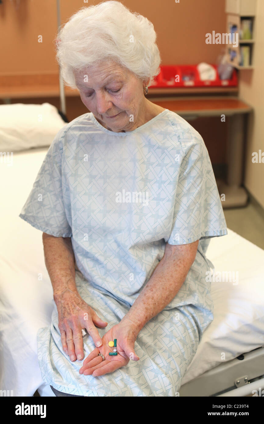 Eine alte Dame in einem Krankenhaus Kleid: sitzen auf einem Bett: blickte auf mehrere Pillen in der Hand Stockfoto