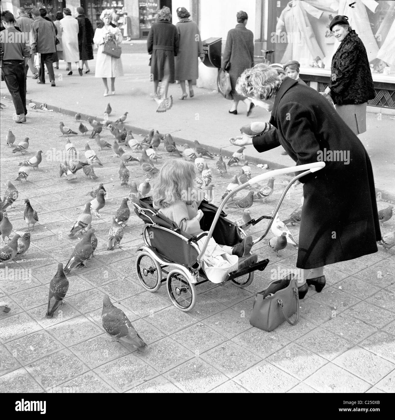 1950er Jahre, eine Dame im Mantel mit einem kleinen Kind in einem Kinderwagen aus der Zeit, die Tauben auf einem gepflasterten Gebiet auf der Avenue des Champs Elysees, Paris, Frankreich fütterte. Stockfoto