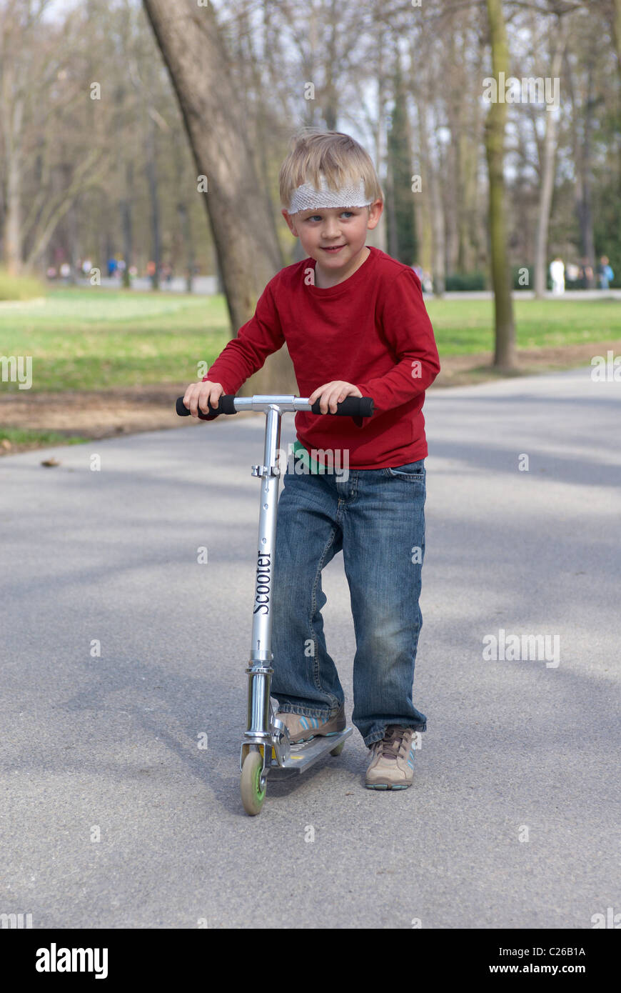 Ein junges Kind blonde junge auf einem Roller im Park mit verletzten Kopf (Capeline Bandage) ohne Schutzhelm Stockfoto
