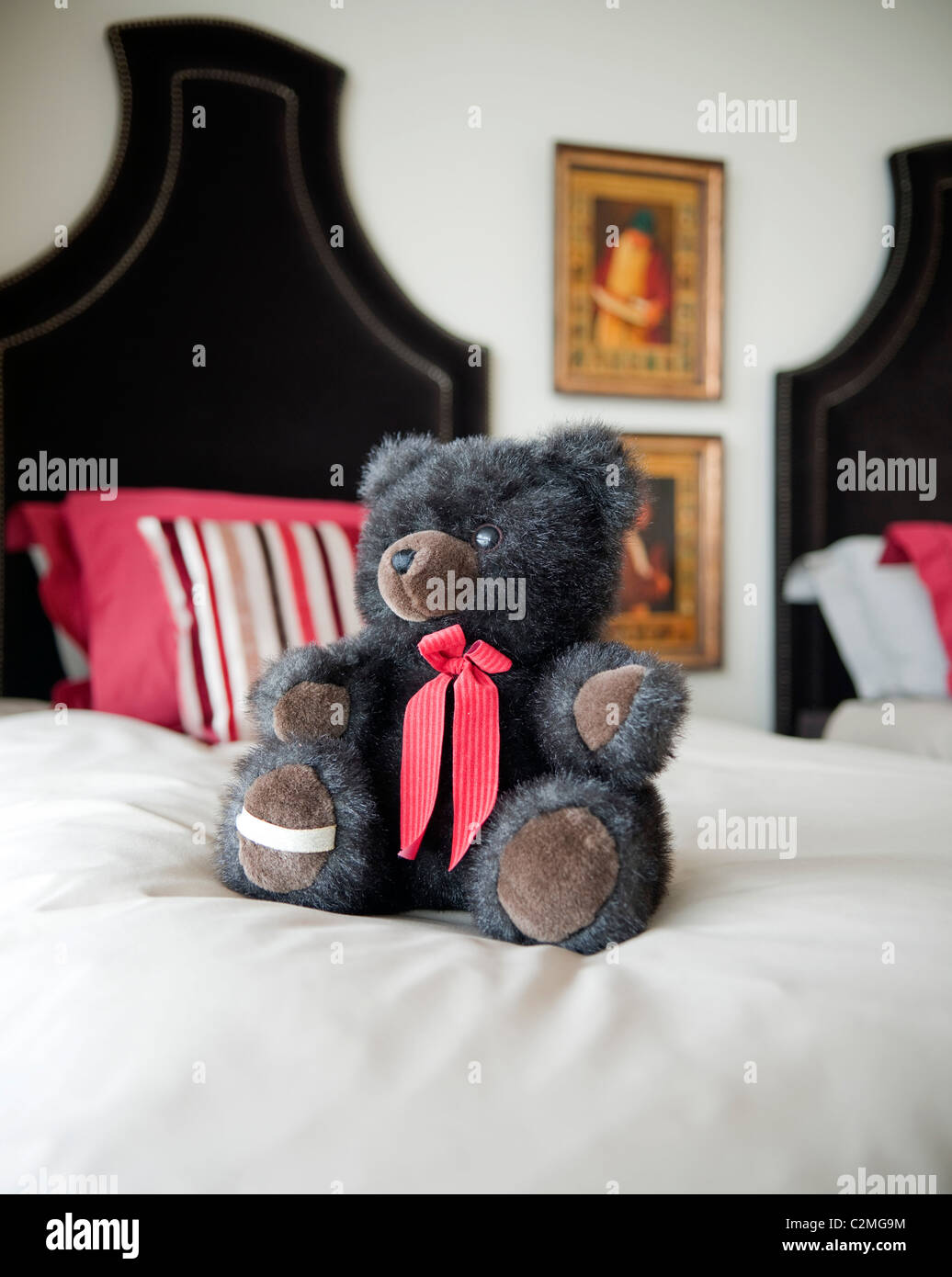 Der Seen-Teddybär mit rotem Band um Hals sitzt auf weißen Bett Stockfoto