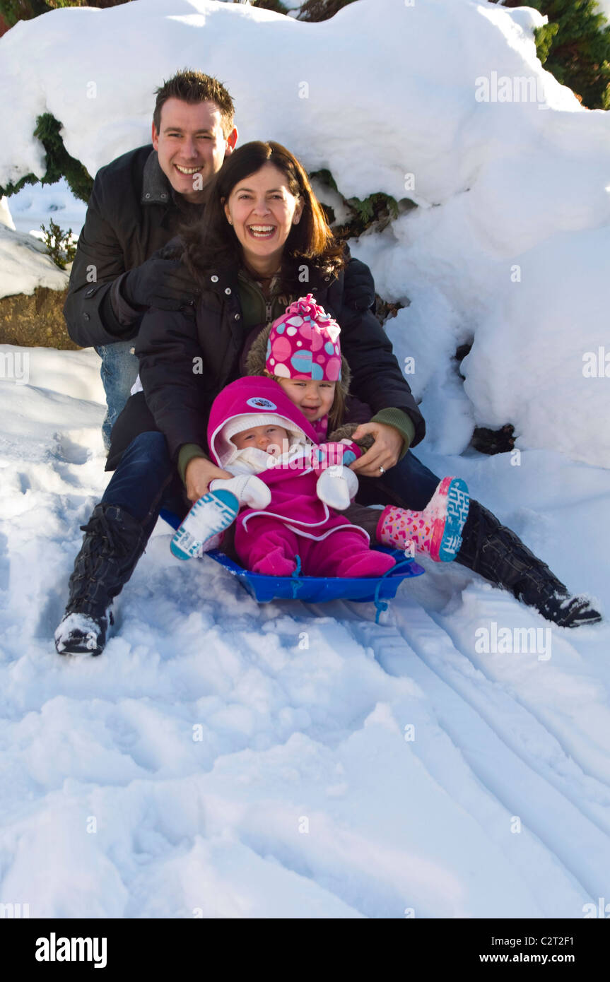 Vertikale Porträt einer jungen Familie, Mama, Papa und zwei junge Mädchen, die alle spielen zusammen auf einem Schlitten oder Schlitten im Schnee Stockfoto