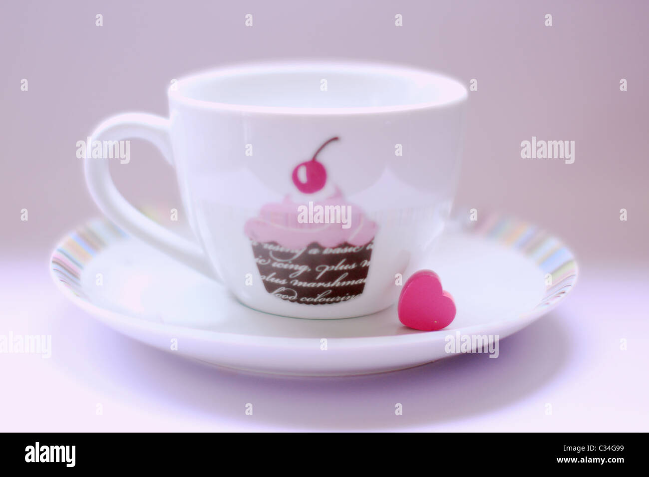 White Teetasse und Untertasse mit einem Cupcake-Design und ein pinkes Herz auf den Teller setzen auf lila Hintergrund Stockfoto