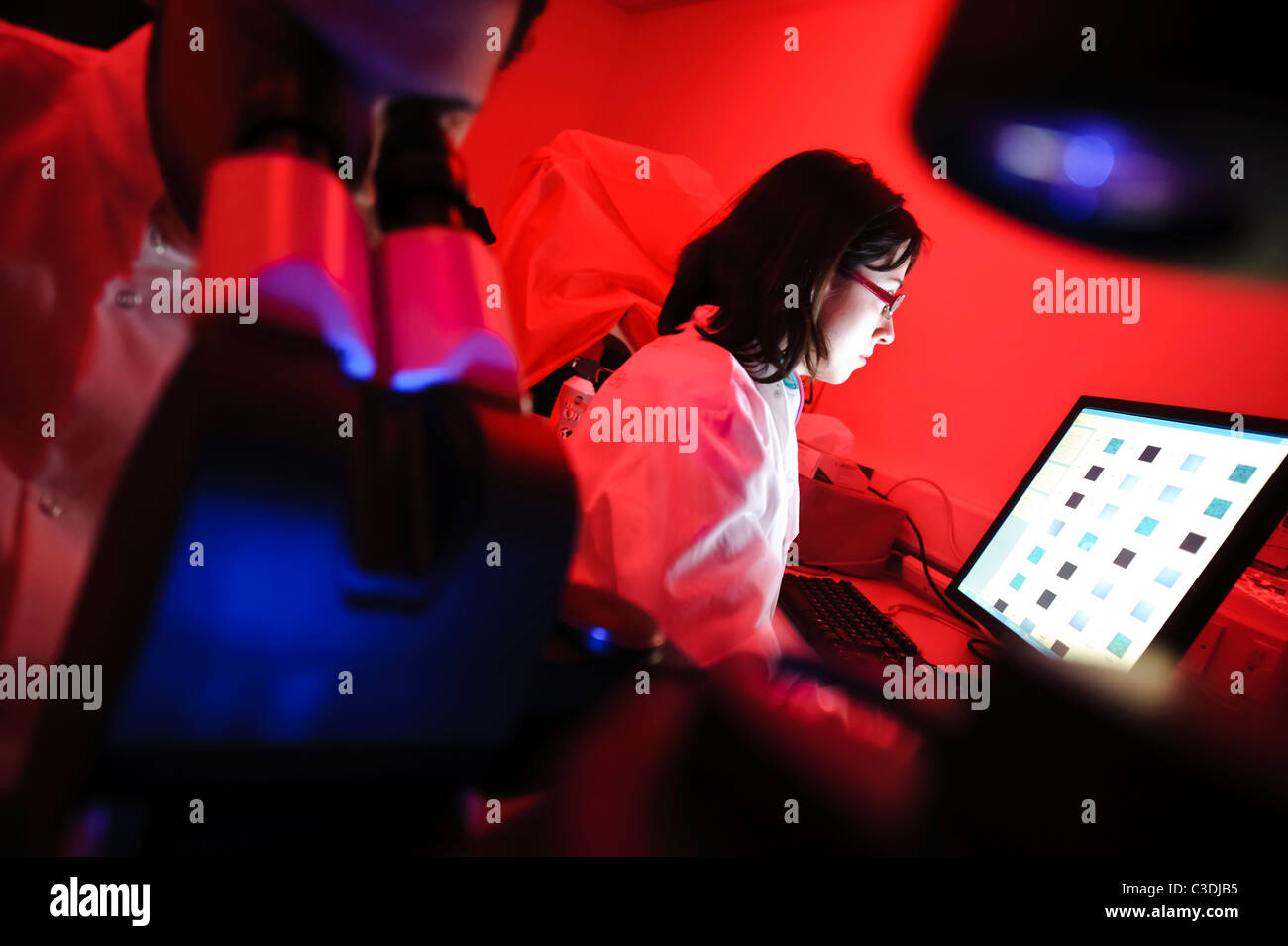 männliche und weibliche Wissenschaftler in Wissenschaft Dunkelkammer mit roter Beleuchtung nach unten Mikroskop mit Bildschirm im Hintergrund Stockfoto
