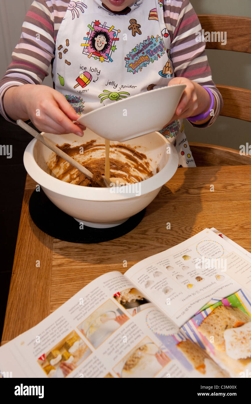Junge Mädchen Kochen, Tracy Becher Schürze tragen, Kuchen backen, Gießen geschlagenen Ei in Schüssel mit Mischung, Kochbuch offen auf dem Tisch - Yorkshire, England. Stockfoto