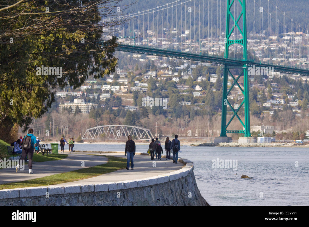 Wanderer und Inlineskater entlang Kaimauer im Stanley Park. Iconic Lions Gate Bridge overhead mit West Vancouver in der Ferne. Stockfoto