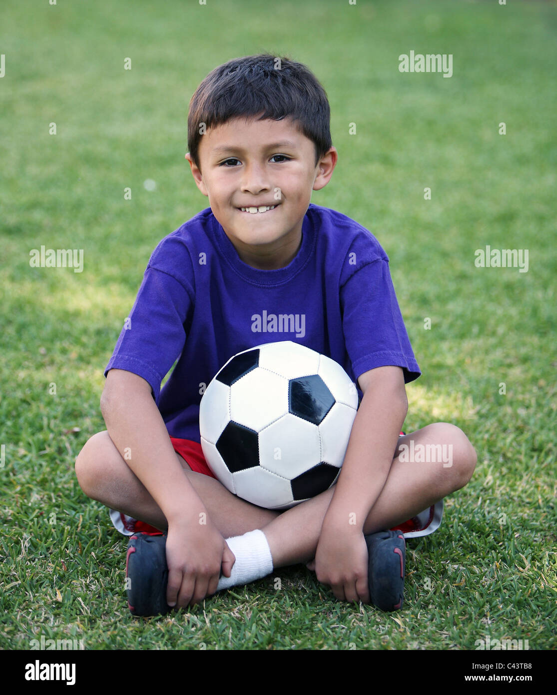 Authentische junge Latino sitzt im Schneidersitz mit Fußball auf Feld Gras Stockfoto
