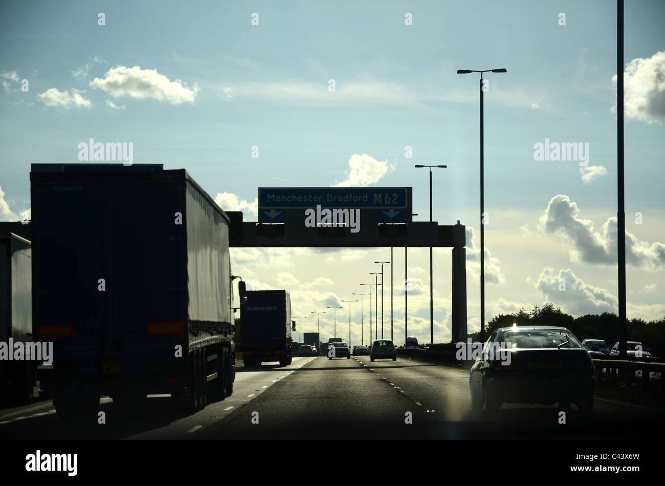 Sicht des Fahrers auf der Autobahn M62 mit Verkehr, Laternenmasten und Beschilderung in der Silhouette gegen einen hellen blauen bewölkten Himmel Stockfoto
