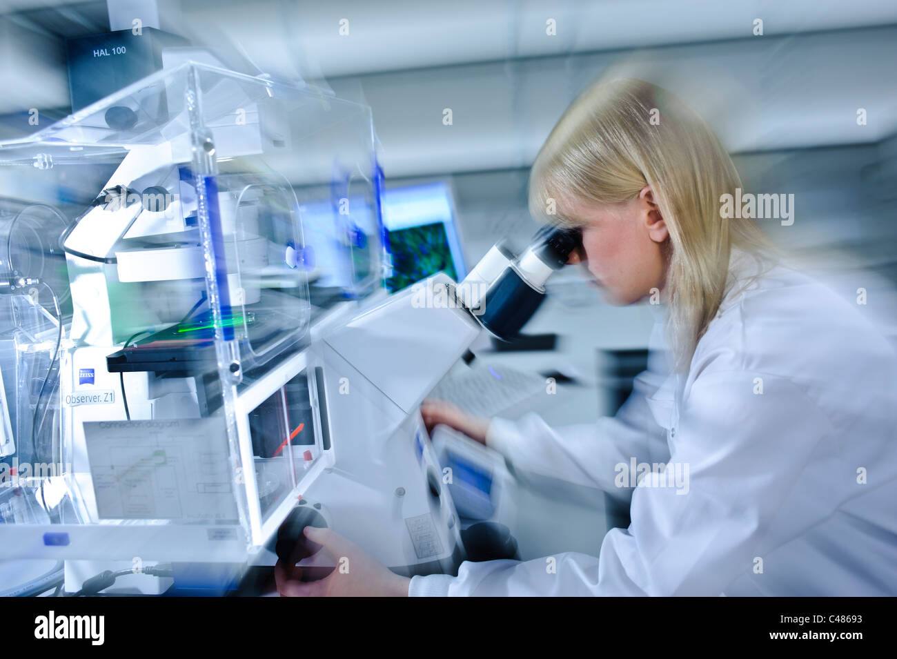 Junge blonde Wissenschaftlerin weißen Lab Coat starken Mikroskop in gut beleuchteten Wissenschaft Labor Zelle Bildern am Computer-Bildschirm Stockfoto