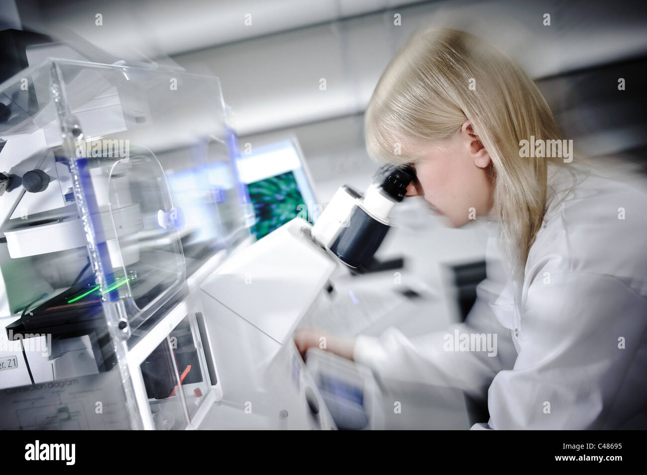 Junge blonde Wissenschaftlerin weißen Lab Coat starken Mikroskop in gut beleuchteten Wissenschaft Labor Zelle Bildern am Computer-Bildschirm Stockfoto