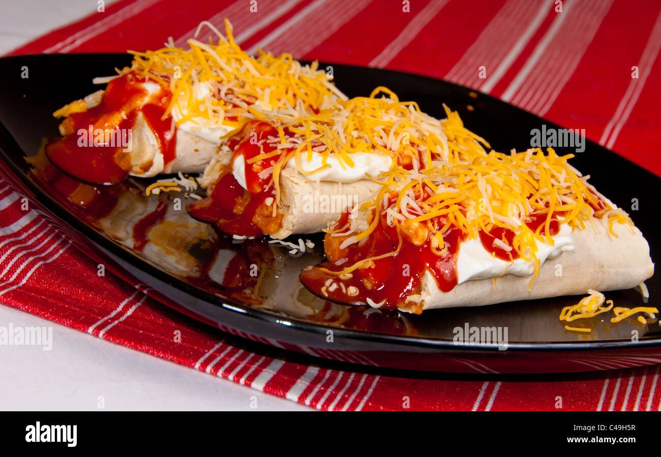 Drei Burritos mit Sauerrahm, Enchiladasoße und geschreddert Käse auf einem schwarzen Schild auf einem roten Tuch bedeckt. Stockfoto