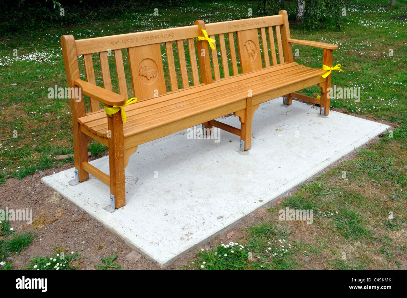 Neue breite Holz öffentlichen Park Sitzbank auf Betonboden Gelbe Band Inschrift & carved Panels als Gedenkstätte auf Village Green Essex England Großbritannien platziert Stockfoto