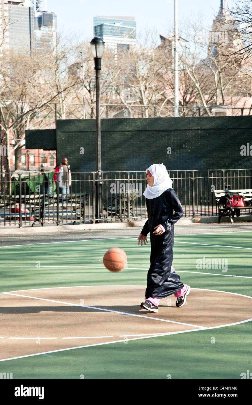 hübsche junges muslimische Mädchen im Kopftuch mit schwarzen Abaya über ihre Jeans spielt mit Fußball in New York City Spielplatz Stockfoto