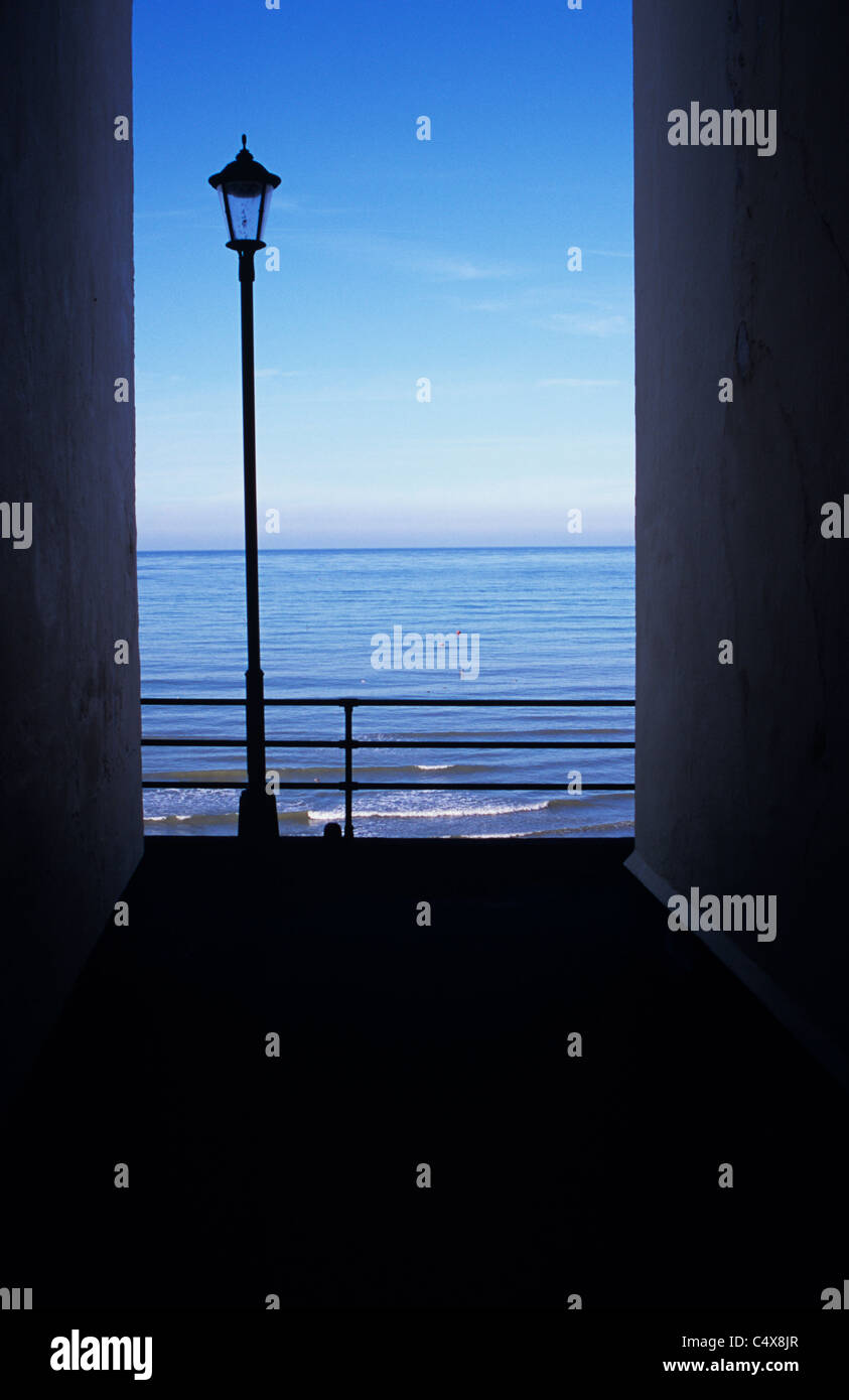 Blick auf ruhige blaue Meer und Himmel mit Geländern und Laternenpfahl Silhouette über ihnen und umrahmt von schattigen Gebäude Stockfoto