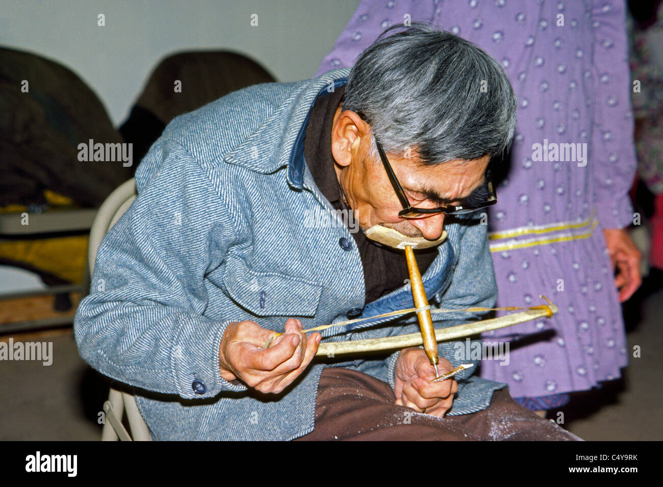 Älteren Eskimo Künstler demonstriert die Verwendung von ein native Hand Tool namens einen Bogen Bohrer um Designs in ein kleines Stück Elfenbein in Nome, Alaska, USA zu machen. Stockfoto